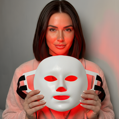 Kvinna håller InfraRelief™ Ansiktsmask med rött LED-ljus aktiverat framför ansiktet mot grå bakgrund