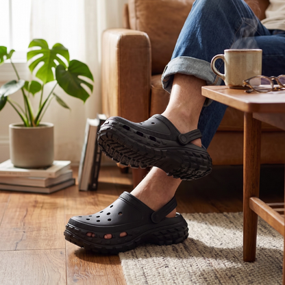 Person sitter i soffa med svarta clogs med terrängssula, lifestyle-bild i hemiljö med trägolv och krukväxter