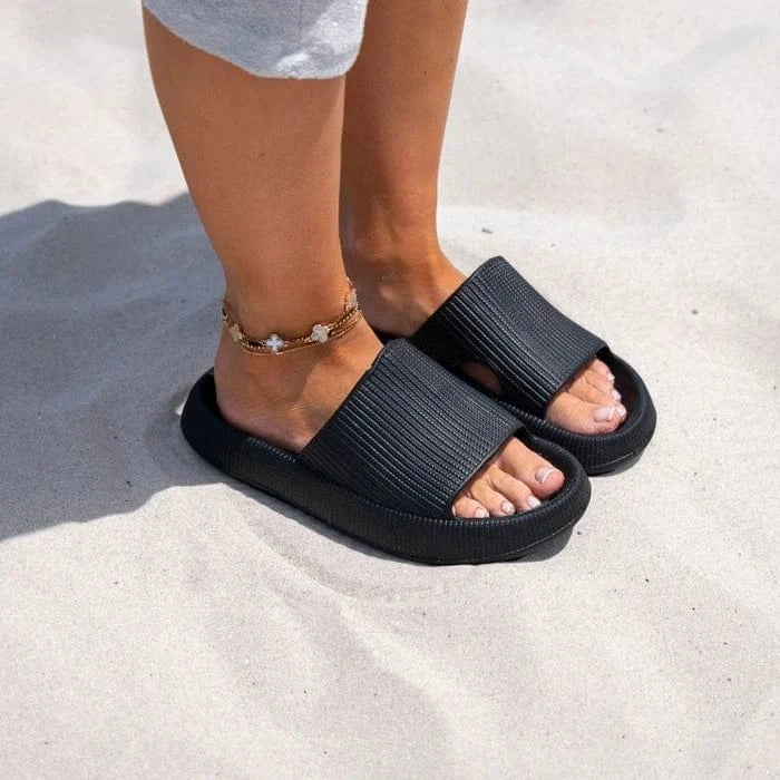 Person bär svarta ergonomiska tofflor på sand – stöddämpande sommartofflor med halkfri sula för strand och utomhusbruk